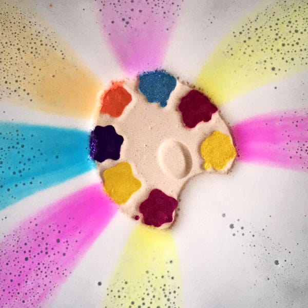 Bath bomb in the shape of a paint palette dissolving in the water, releasing colorful swirls.